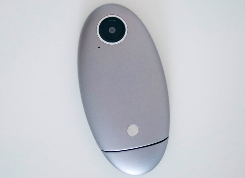 A silver, oval-shaped electronic device with a camera lens at one end, lying on a white surface.