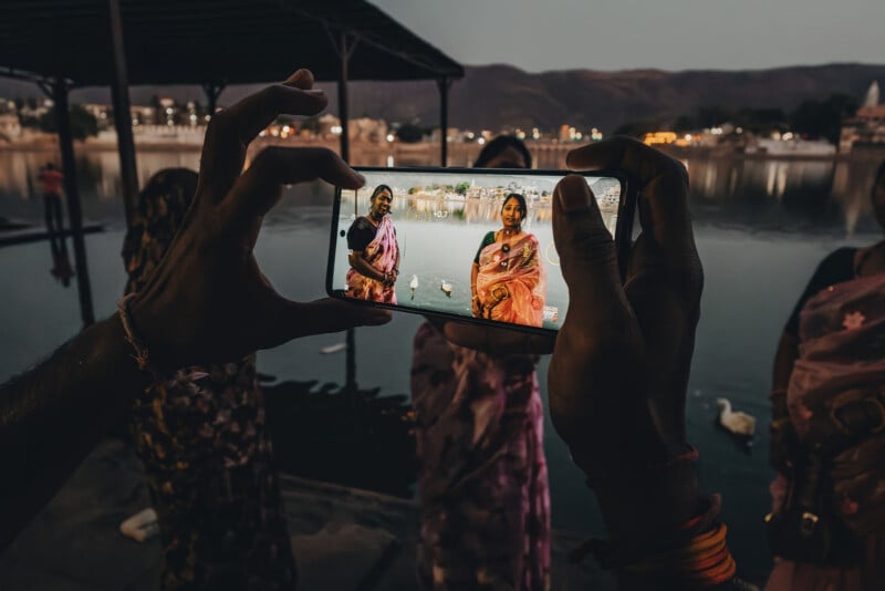 A person holds a phone, capturing two women in colorful sarees standing by a lakeside at dusk, with city lights and mountains in the background. The phone screen shows the women clearly, while the background is darker.