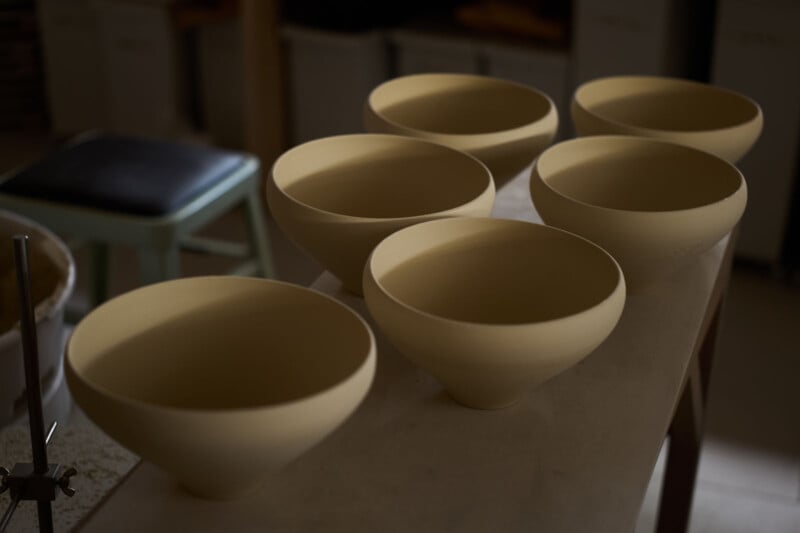 Six unglazed, light clay bowls with wide rims and tapered bases are arranged in two rows on a white table in a softly lit pottery studio. A stool and shelves are visible in the background.